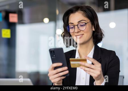 Young businesswoman holds smartphone, engages in online shopping with credit card in modern office. Highlights digital transaction, financial technology, business innovation, and mobile security. Stock Photo