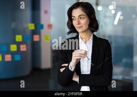 Confident businesswoman in office setting holding glasses, dressed in professional attire. Concept of leadership, contemplation in work environment. Vibrant sticky notes add creative touch. Stock Photo