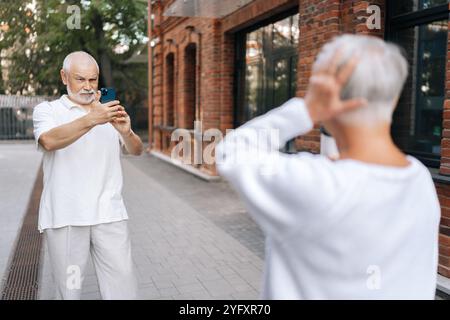 Rear view of happy senior couple capturing moments in city street with smartphone on sunny summer day, enjoying retirement, exploring urban landscapes Stock Photo