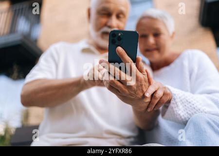 Selective focus shot of elderly married couple using smartphone, exploring apps, staying connected with world, embracing digital age in retirement. Stock Photo