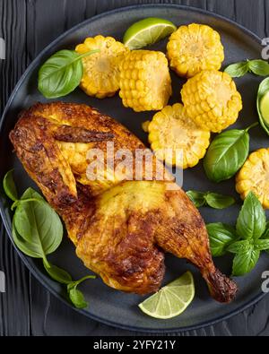 half of roasted chicken with corn cobs coins and fresh basil leaves on plate on black wooden table, close-up Stock Photo