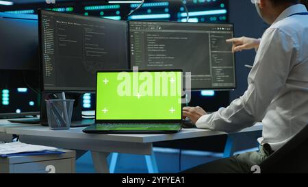 Zoom in on green screen laptop used by data center worker troubleshooting issues. Engineer uses chroma key notebook in server hub, establishing performance benchmarks, camera A Stock Photo