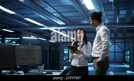 Team of technicians working on tablets in data center used for managing gear energy consumption. Teamworking colleagues using devices in workspace used for automation in server farm, camera B Stock Photo