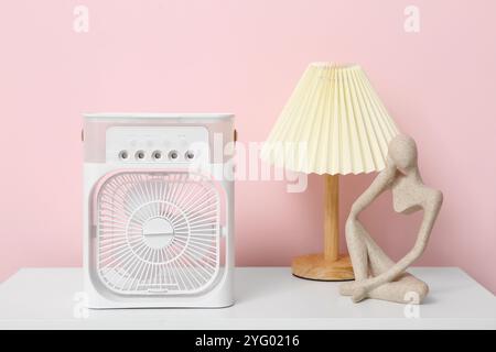 Portable table fan with statuette and lamp on shelf near pink wall Stock Photo