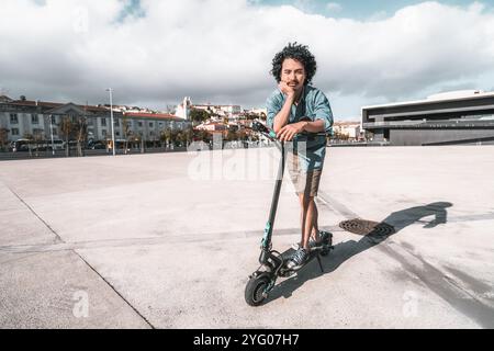 Man with curly hair in a casual outfit stands thoughtfully next to an electric scooter on a large, empty urban plaza. He leans on the scooter with one Stock Photo
