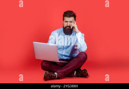 Man working on laptop. Beard man sitting on the floor with and using laptop computer on red background. Holding laptop computer. Businessman using his Stock Photo