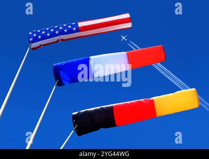 three windsocks in strong winds, in the colours of the national flags of the USA, France and Germany against a blue background and a passenger plane Stock Photo