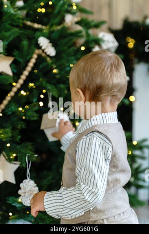 A little boy in a shirt and vest stands facing a Christmas tree decorated Stock Photo