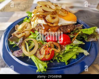 Close up of a decorated blue dinner plate filled with a Hamburger fried egg fried onion rings tomatoes and lettuce with a topping Stock Photo