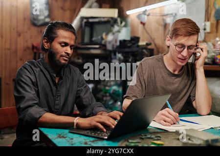 Young technician with smartphone by ear taking notes and phoning client while his colleague typing on laptop keyboard Stock Photo