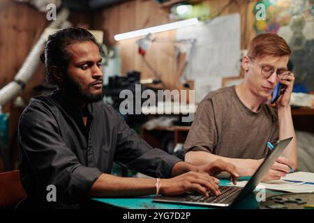 Young technician typing on laptop keyboard against colleague taking notes and talking to client on mobile phone Stock Photo