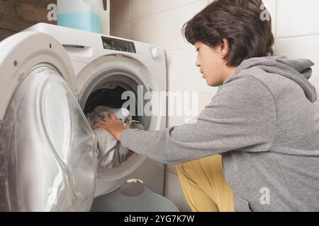 A child puts the dirty clothes in the washing machine. He is wearing a gray hoodie Stock Photo