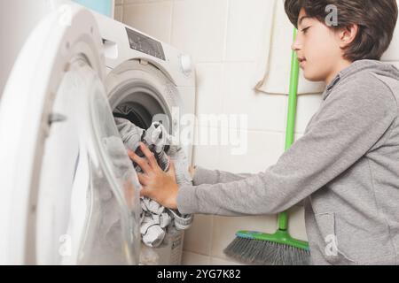 A young boy is putting clothes in a washing machine. The boy is wearing a gray hoodie Stock Photo