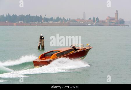 A classic Venetian water taxi speeds across the lagoon, leaving a trail of waves, with the serene backdrop of Venice’s historic architecture. Stock Photo