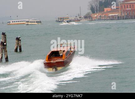 A classic Venetian water taxi speeds across the lagoon, leaving a trail of waves, with the serene backdrop of Venice’s historic architecture. Stock Photo