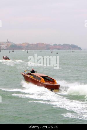 A classic Venetian water taxi speeds across the lagoon, leaving a trail of waves, with the serene backdrop of Venice’s historic architecture. Stock Photo