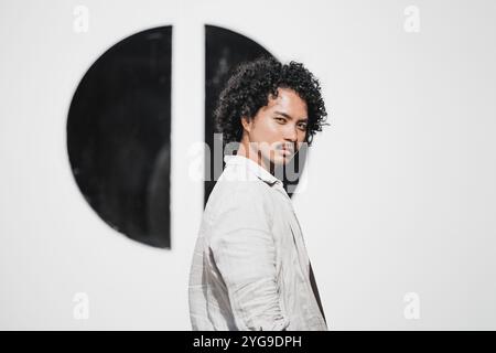 Portrait of a young man with curly hair and a mustache, wearing a white linen shirt, looking confidently at the camera, standing in front of a minimal Stock Photo
