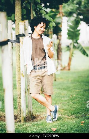 Young man with curly hair wearing a casual outfit, standing outdoors in a park. He's leaning against a tree, holding a smartphone with a smile, dresse Stock Photo