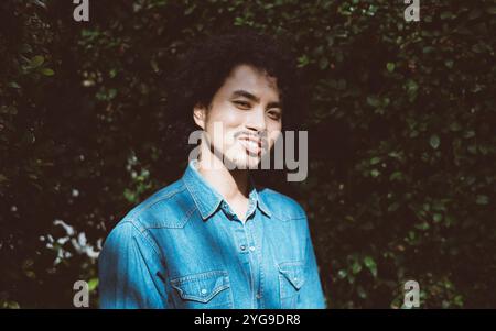 Portrait of a young man with curly hair and a goatee, wearing a blue denim shirt, standing outdoors against a lush green bush background. Sunlight cas Stock Photo
