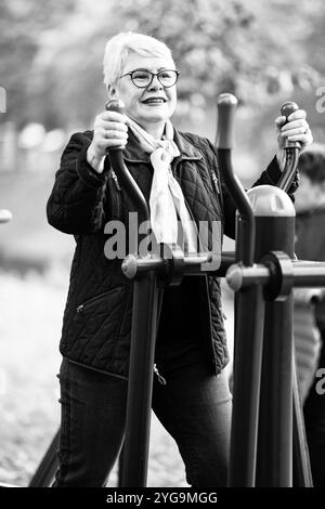 An older woman performs a workout on an exercise simulator in a park, dressed in blue clothing. With a casual outfit and glasses, she demonstrates act Stock Photo