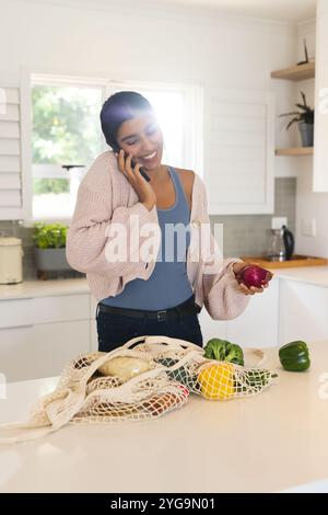 Smiling woman in kitchen talking on phone while holding fresh vegetables, at home Stock Photo