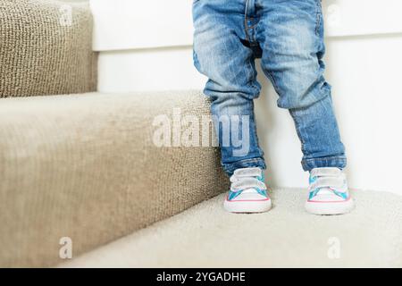 Toddler standing at the edge of carpeted stairs wearing jeans and colourful sneakers Stock Photo