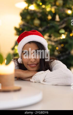 Asian woman wearing Santa hat smiling by Christmas tree, enjoying festive season, at home Stock Photo