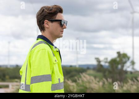 Smart young engineer technician male worker standing side view closeup at Wind Turbines clean power generator field background. Stock Photo