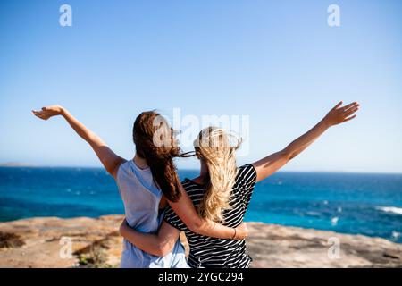 Two young women friends, blonde and brunette, enjoy their summer holiday, embracing with hands in the air and wind in their hair as they stand on a cl Stock Photo