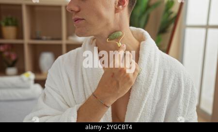 Young man in a spa wearing a white robe and using a jade roller on his neck in a wellness room Stock Photo