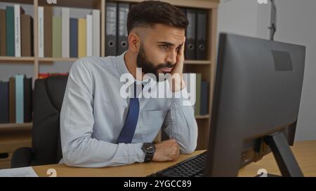 Hispanic man with a beard in an office, seated at a desk, looking at a computer screen, wearing a shirt and tie, appearing thoughtful or concerned, wi Stock Photo