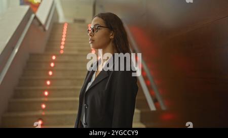 Young woman in a professional setting, standing indoors near illuminated stairs in a lobby or corridor, showcasing a confident and focused demeanor Stock Photo
