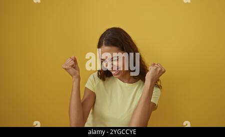 Young woman celebrating with a joyful expression over an isolated yellow background wall, showcasing beautiful hispanic and african american features Stock Photo