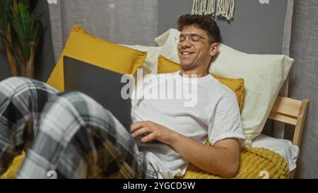 Handsome young hispanic man in a bedroom at home, lying on a bed with yellow pillows, wearing glasses, and using a laptop. Stock Photo