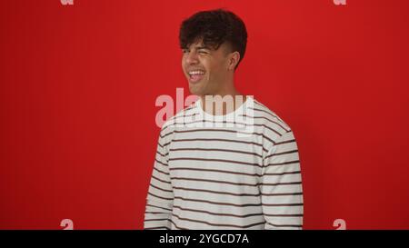 Young hispanic man winking in a striped shirt against a vibrant red background, capturing a playful and charismatic moment. Stock Photo