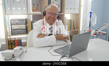 Elderly, grey-haired, caucasian man in doctor attire at the hospital clinic office, smiling while wearing a headset and using a laptop. Stock Photo