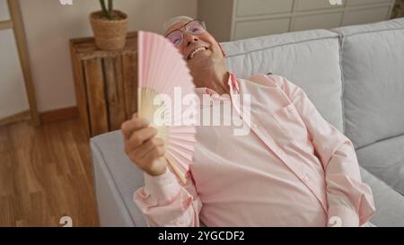 A senior caucasian man relaxing indoors at home, sitting on a sofa holding a pink hand fan, wearing glasses and a pink shirt, in a cozy living room se Stock Photo