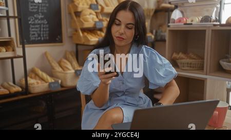 Woman using smartphone while sitting in bakery with bread shelves in background and laptop on table Stock Photo