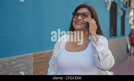 A beautiful mature hispanic woman with short hair and glasses talks on her phone while smiling joyfully on an urban street on a sunny day. Stock Photo