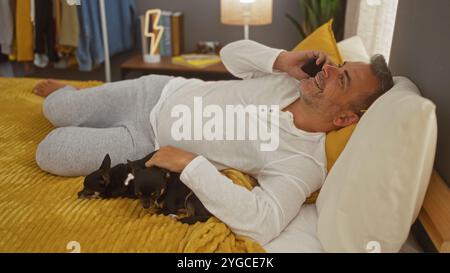 Middle-aged hispanic man relaxing on bed in bedroom with two chihuahuas while talking on phone. Stock Photo