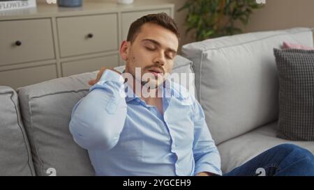 Handsome young hispanic man in a blue shirt with a beard sits in a modern living room, appearing tired and touching his neck, suggesting possible neck Stock Photo