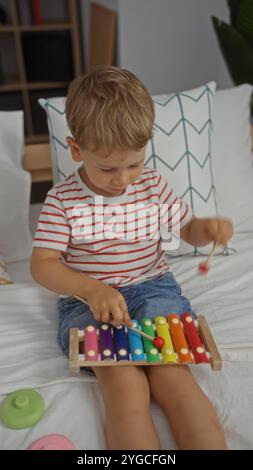 Toddler boy playing xylophone on bed in bright bedroom with striped shirt and denim shorts Stock Photo