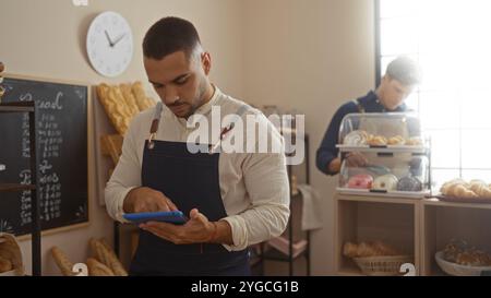 Men working in a bakery, one operating a tablet while the other arranges pastry, showcasing a modern indoor shop environment with various bread and pa Stock Photo