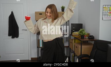A beautiful young blonde woman dances joyfully in an office workplace, holding a red cup, surrounded by shelves, binders, and office supplies, creatin Stock Photo