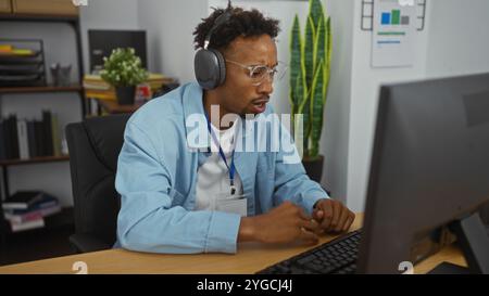 Young african american man with beard and glasses wearing headphones and working intently on a computer in a modern office interior with shelves and p Stock Photo