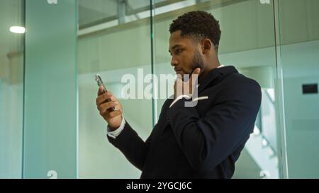 Young man in a suit checking his phone thoughtfully while standing in a modern office setting, surrounded by glass walls and bright lighting. Stock Photo