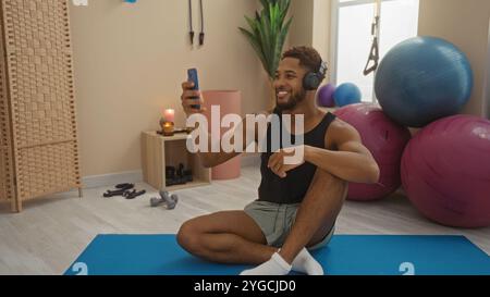 Young man exercising indoors sitting on yoga mat in gym smiling wearing headphones using phone Stock Photo