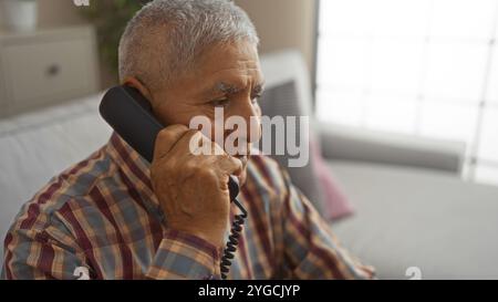 Middle-aged hispanic man with grey hair talking on a landline phone in a cozy living room. Stock Photo