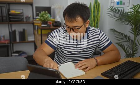 Young, asian man working intensely at his desk in a modern office, concentrating on writing notes while reviewing information on a tablet, surrounded Stock Photo