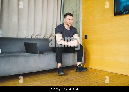 Handsome young man in t-shirt sitting on sofa and drinking coffee, working remotely during startup as a freelancer, looking at laptop and smiling Stock Photo